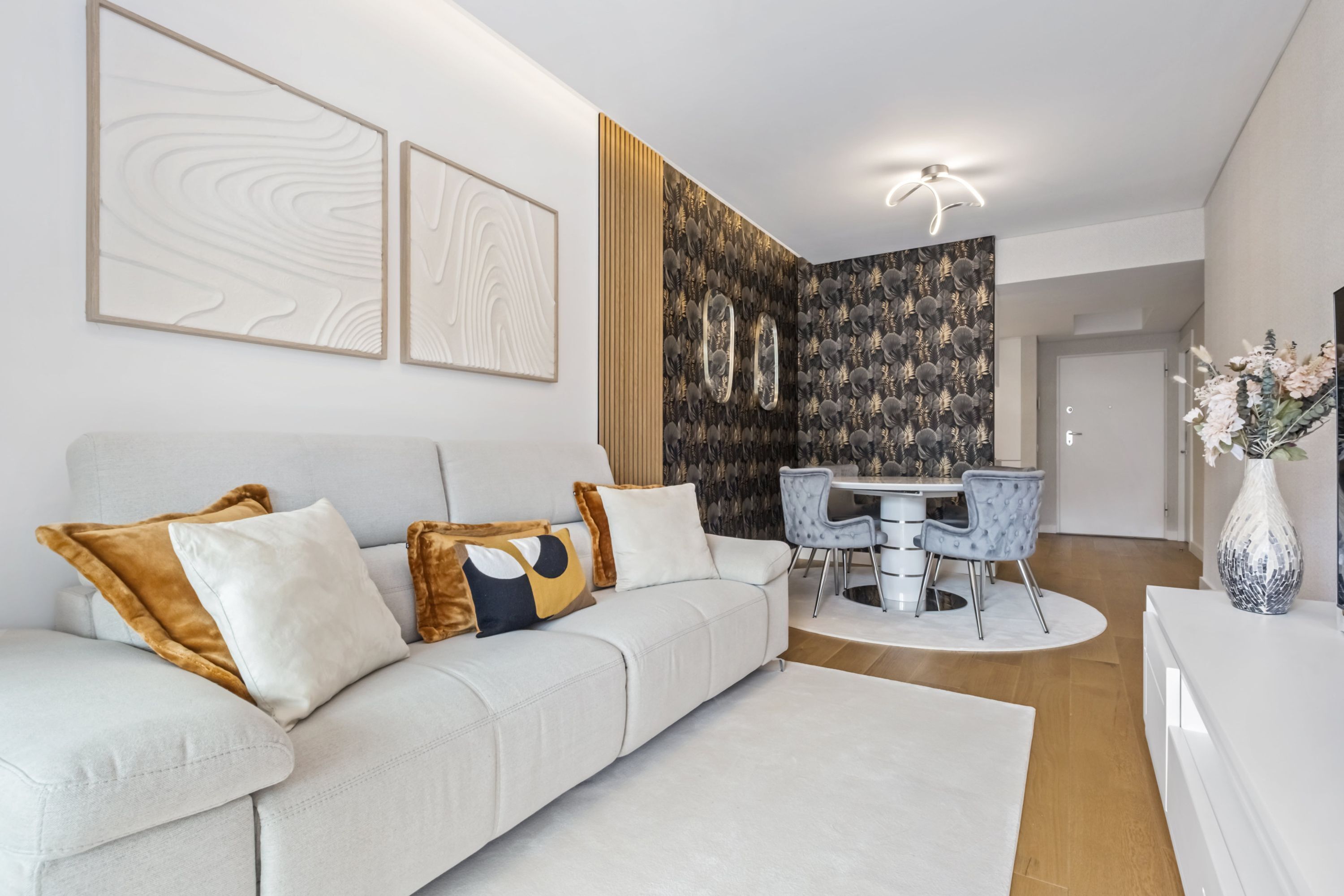 Un salón moderno con un sofá blanco, una mesa de comedor con sillas y una pared decorativa con motivos abstractos.