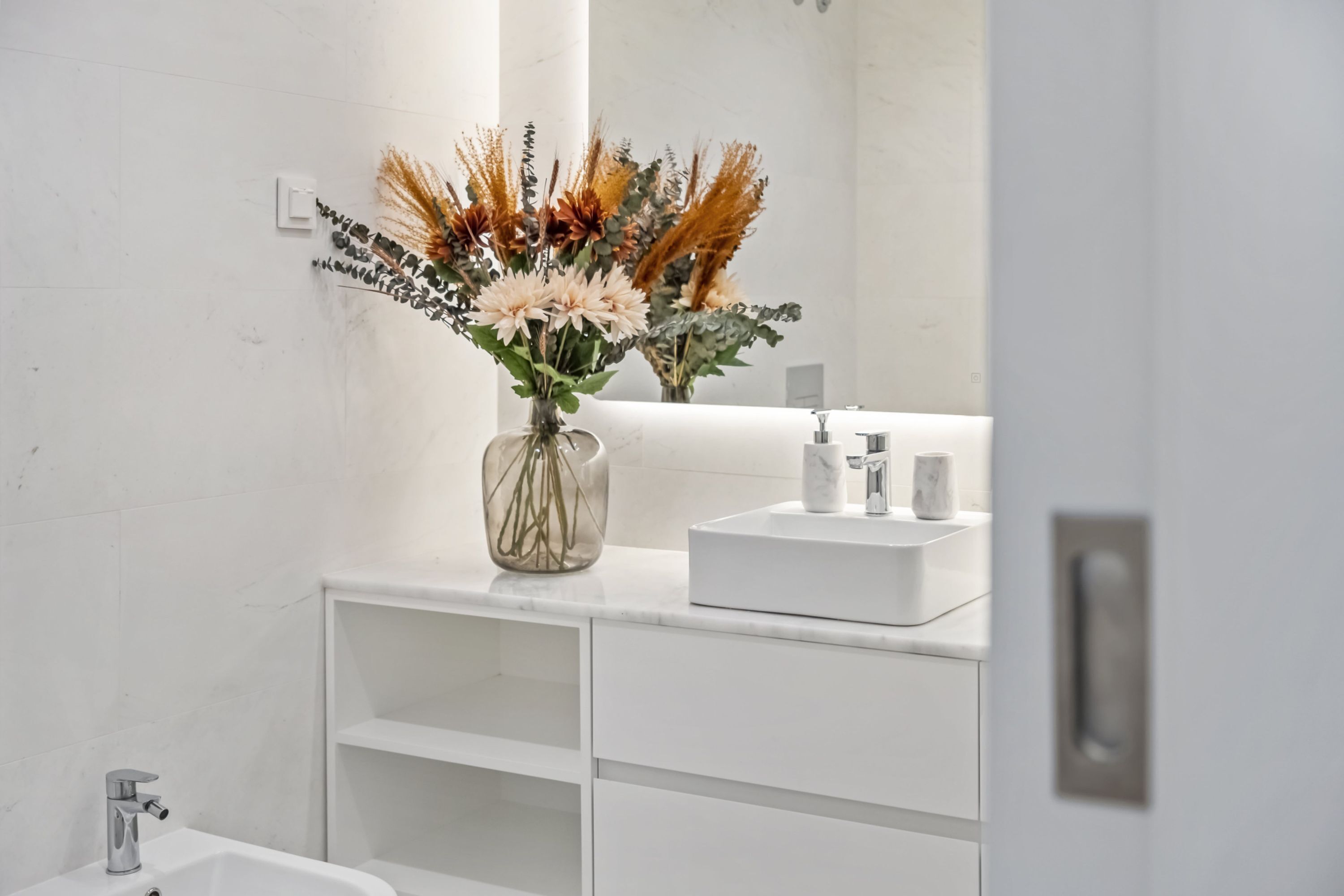 A bathroom with a white sink, mirror, and a vase with dried flowers.