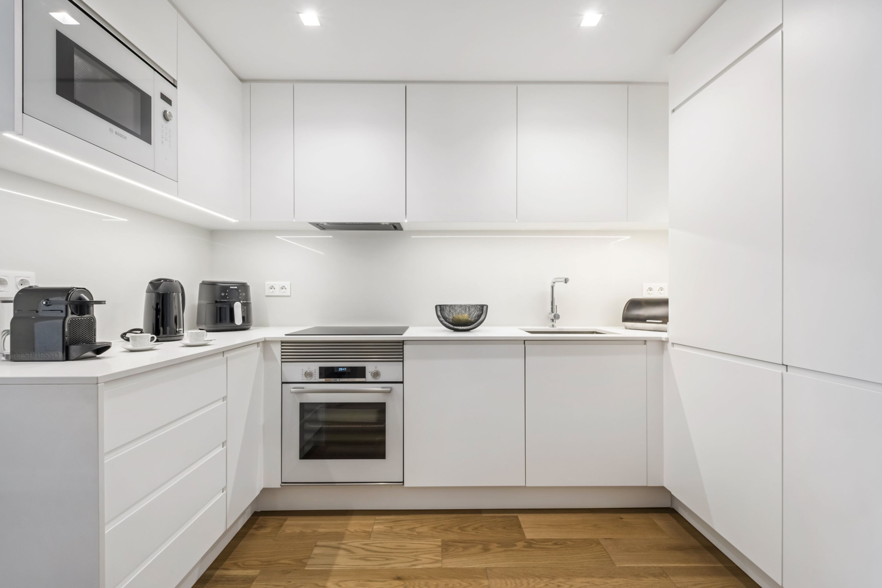 A modern kitchen with white cabinetry, a microwave, a coffee maker, a kettle, a sink, and a stove.