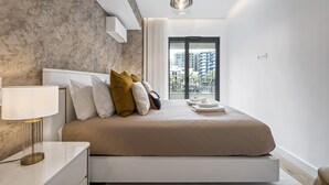 Quarto moderno com cama grande, mesa de cabeceira, luminária e vista para prédios pela janela.