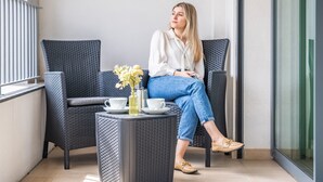 Uma mulher sentada em uma cadeira de vime escura em um quarto moderno, com uma pequena mesa e um vaso de flores.