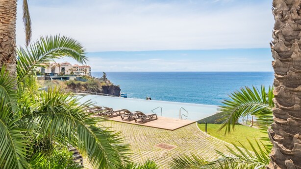 Vista da piscina com espreguiçadeiras, uma visão clara do oceano e um edifício ao fundo.