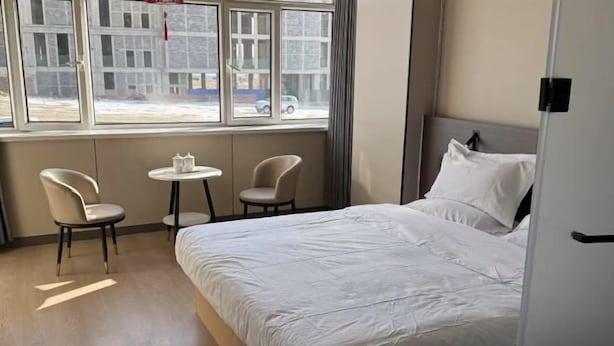 Habitación de hotel con cama, dos sillas, una mesita y ventana con vistas a edificios y coches.
