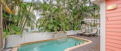 A swimming pool with a wooden deck, surrounded by lush greenery and a white fence.