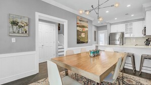 A modern kitchen with a stainless steel refrigerator, a dining area with a wooden table and chairs, and a staircase leading to another level. - 125 Grand Pavillion by Avantstay Steps From Sand (Isle of Palms)