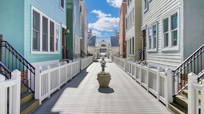 A wooden boardwalk leading to a central archway between colorful buildings. - 125 Grand Pavillion by Avantstay Steps From Sand (Isle of Palms)