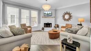 A living room with a fireplace, a television mounted above it, and a chandelier. - 125 Grand Pavillion by Avantstay Steps From Sand (Isle of Palms)