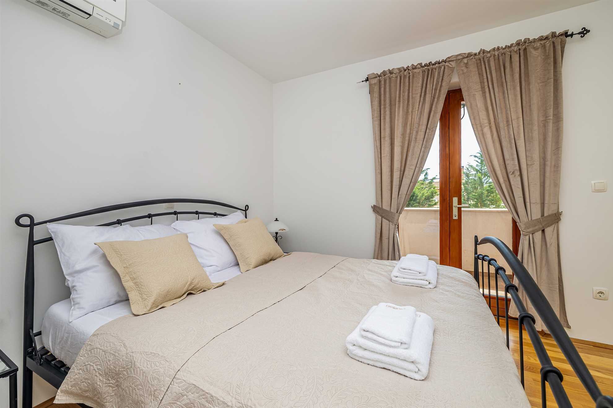 A neatly made bed with a beige bedspread and pillows, a window with curtains, and a balcony railing.
