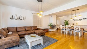 A spacious living room with a brown sectional sofa, a white coffee table, and a fireplace. There is a dining area with a wooden table and chairs, and a kitchen visible in the background.