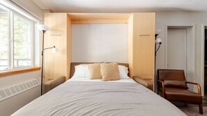 Un dormitorio con cama, una silla, una mesita de noche, una lámpara y una ventana.