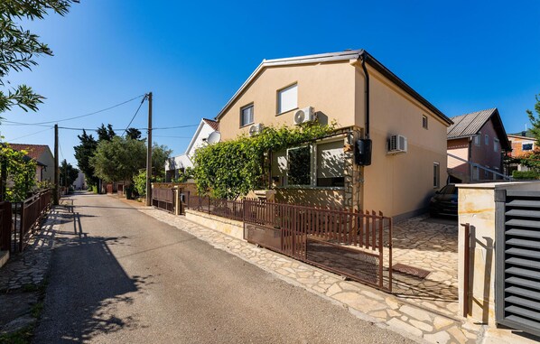A residential street with a beige house featuring a stone facade, a metal fence, and a car parked in the driveway.