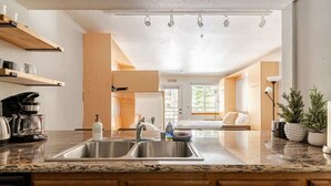 Eine moderne Küche mit einer Spüle, Holzschränken und einer Arbeitsplatte. Über der Spüle befinden sich Regale und rechts eine Zimmerpflanze.