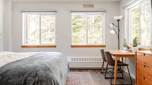 Un dormitorio con cama, escritorio, silla, cómoda y dos ventanas con vistas a árboles.