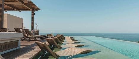 Un espace aménagé au bord de la piscine, avec des chaises longues et une pergola en bois offrant une vue sur l’océan.