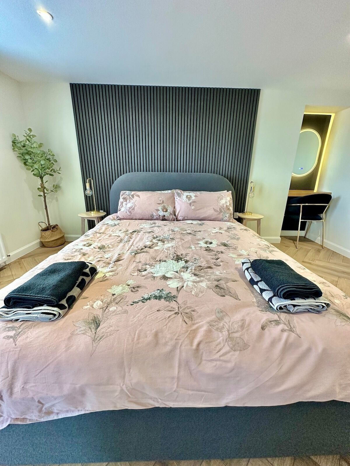 A neatly made bed with a floral bedspread and two rolled towels, a bedside table with a lamp, a small potted plant, and a mirror on the wall.