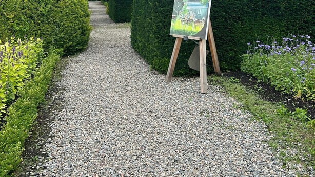 A gravel pathway leading to a traditional stone building with a well-maintained garden and a painting on an easel.
