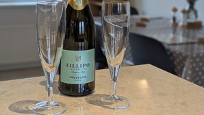 A bottle of FILIPPO Prosecco and two glasses on a marble table in a room with a TV and a window with pink flowers.