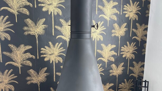 A modern fireplace with a black chimney, set against a wall with a gold palm tree wallpaper.