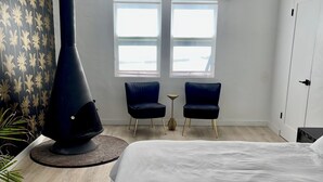 A bedroom with a large bed, a black heater, two chairs, a small table, and a window with an air conditioner.