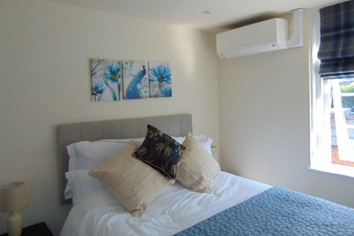 A bedroom with a bed, a nightstand, a wall-mounted air conditioner, and a window with curtains.
