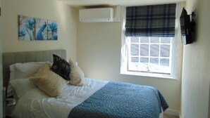 A bedroom with a bed, a window with blinds, a wall-mounted air conditioner, and a painting on the wall. - The Three Crowns (Loughborough)