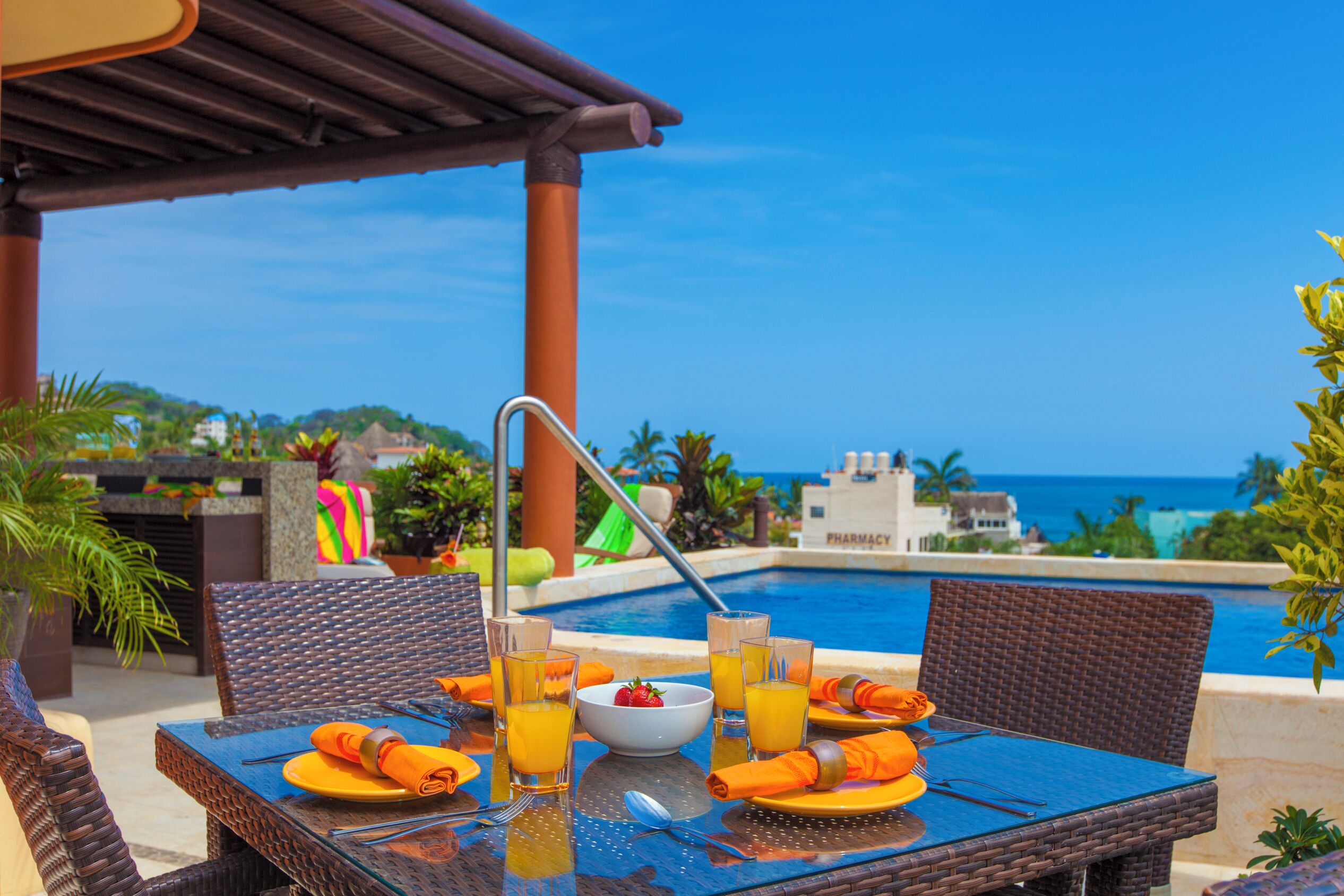 A dining table with a view of a pool and ocean, surrounded by wicker chairs and orange napkins.