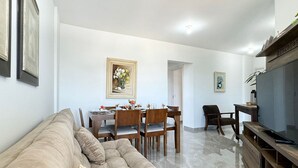 Una sala de estar con un sofá beige, una mesa de comedor con sillas, un televisor y obras de arte enmarcadas en las paredes.