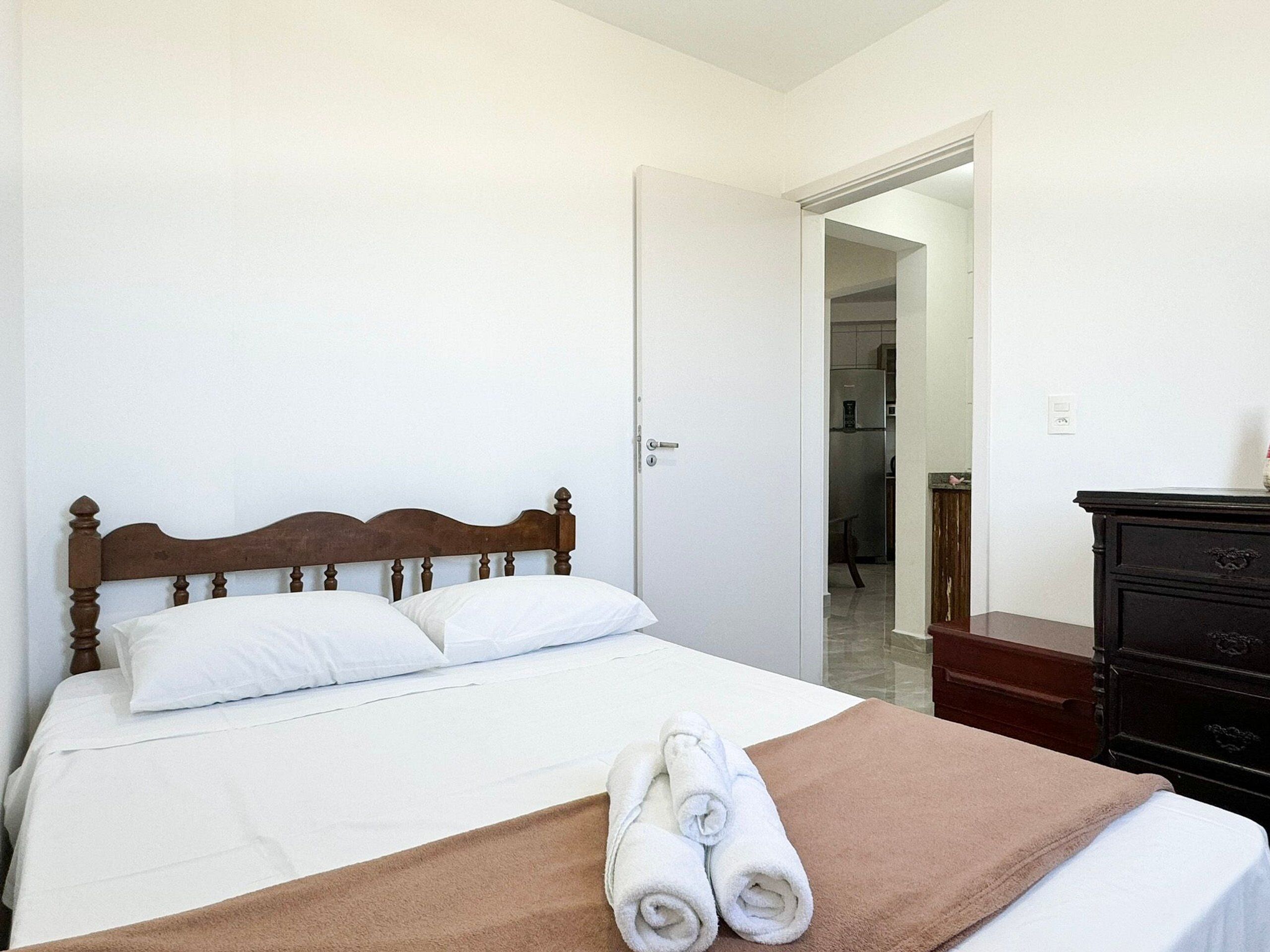Cama bem arrumada com cabeceira de madeira, lençóis brancos e manta marrom. Um aparador de madeira escura está à direita.