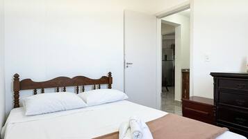 Cama bem arrumada com cabeceira de madeira, lençóis brancos e manta marrom. Um aparador de madeira escura está à direita.
