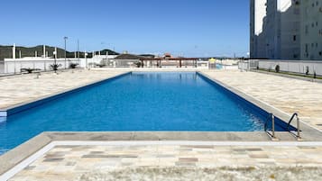 Piscina retangular com um deck ladrilhado ao redor, uma escada de metal e um céu azul claro ao fundo.
