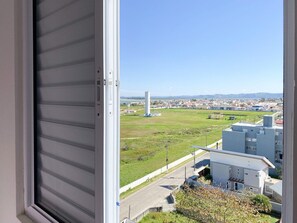 A window view of a residential area with a clear sky, a large industrial structure, and a road with a fence. - Resid. Clube Mar Grosso-Laguna - LAG801 (Laguna)