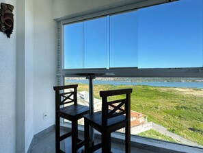 Ein Balkon mit zwei Holzstühlen, ein Fenster mit Blick auf ein Gewässer und Grünflächen.