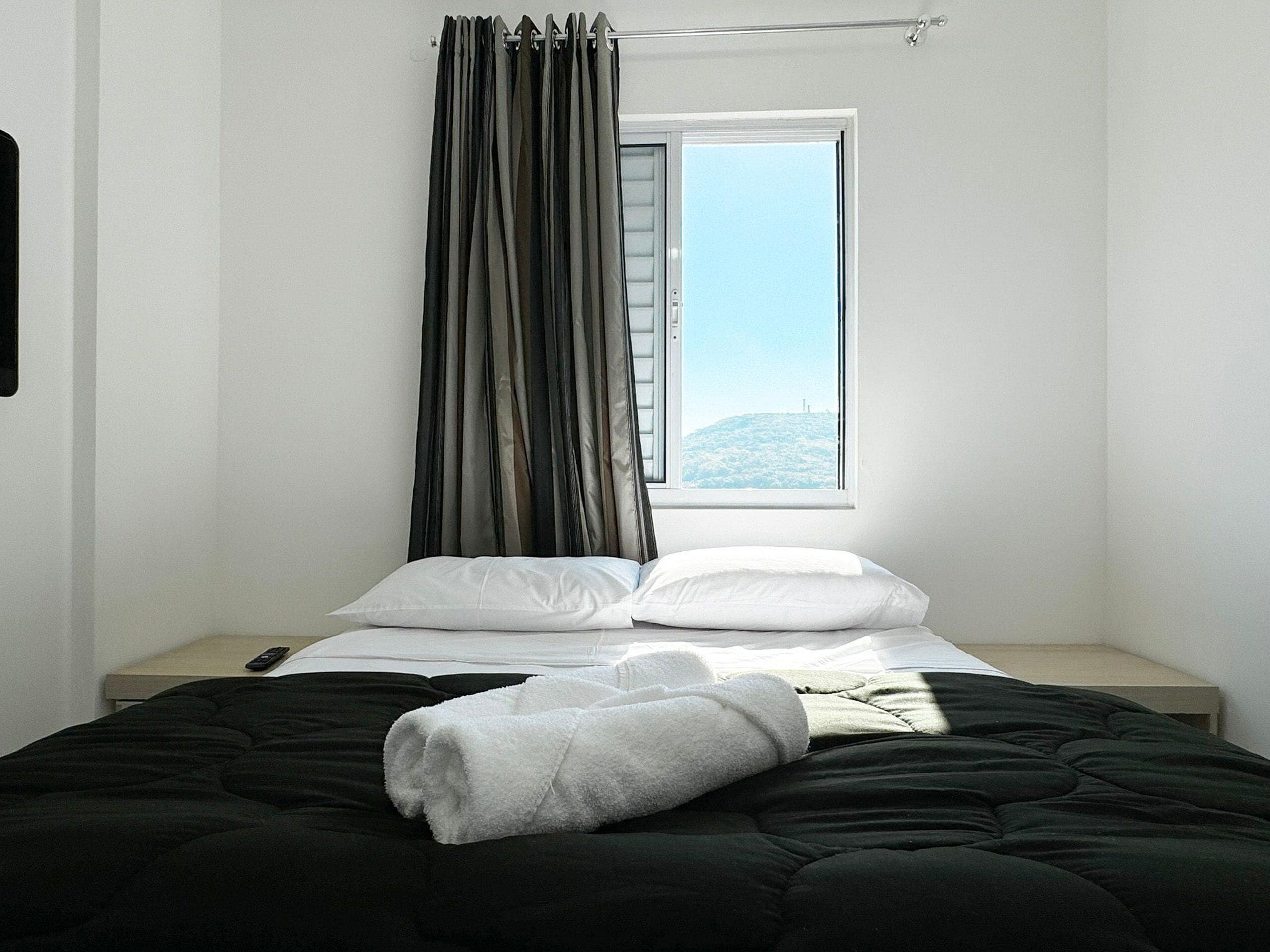 Un dormitorio con cama, almohadas blancas, edredón negro, toalla blanca y una ventana con cortinas.