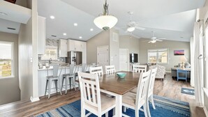 A spacious kitchen with white cabinets, a dining area with a wooden table and white chairs, and a living room with a sofa and a TV. - The Beachcomber by Topsail Realty (Holly Ridge)