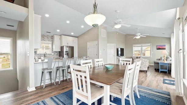 A spacious kitchen with white cabinets, a dining area with a wooden table and white chairs, and a living room with a sofa and a TV. - The Beachcomber by Topsail Realty (Holly Ridge)