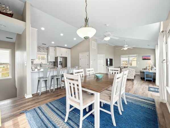 A spacious kitchen with white cabinets, a dining area with a wooden table and white chairs, and a living room with a sofa and a TV. - The Beachcomber by Topsail Realty (Holly Ridge)