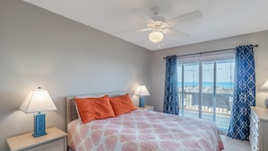 A bedroom with a bed, two bedside tables with lamps, a ceiling fan, and a view of the beach through a large window. - The Beachcomber by Topsail Realty (Holly Ridge)