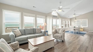 A spacious living room with a large window overlooking the ocean, a ceiling fan, and a dining area with a view. - The Beachcomber by Topsail Realty (Holly Ridge)