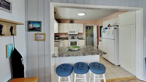 A kitchen with a granite countertop, white cabinets, a microwave, and a refrigerator. There are three blue barstools and a wooden table. - Costic by Topsail Realty (Holly Ridge)