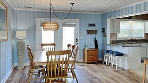 A dining area with a wooden table and chairs, a hanging light fixture, and a kitchen area with stools and a counter. - Costic by Topsail Realty (Holly Ridge)