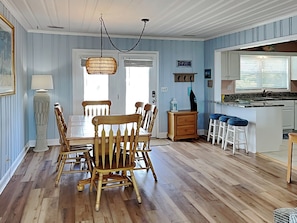 A dining area with a wooden table and chairs, a hanging light fixture, and a kitchen area with stools and a counter. - Costic by Topsail Realty (Holly Ridge)
