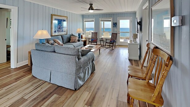 A living room with a sofa, chairs, wooden flooring, a ceiling fan, and a view of the ocean through a door. - Costic by Topsail Realty (Holly Ridge)
