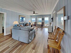 Un salon comprenant un canapé, des fauteuils, un plancher en bois, un ventilateur de plafond et une vue sur l’océan par une porte.