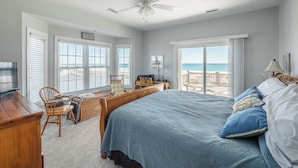Un dormitorio con una cama grande, un escritorio y una silla. Hay ventanas con persianas, un ventilador de techo y vistas a la playa.