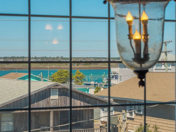 A view through a window with a grid pattern, overlooking a body of water, a dock, and buildings with a wooden structure and a utility pole. - Serendipity by Topsail Realty (Holly Ridge)