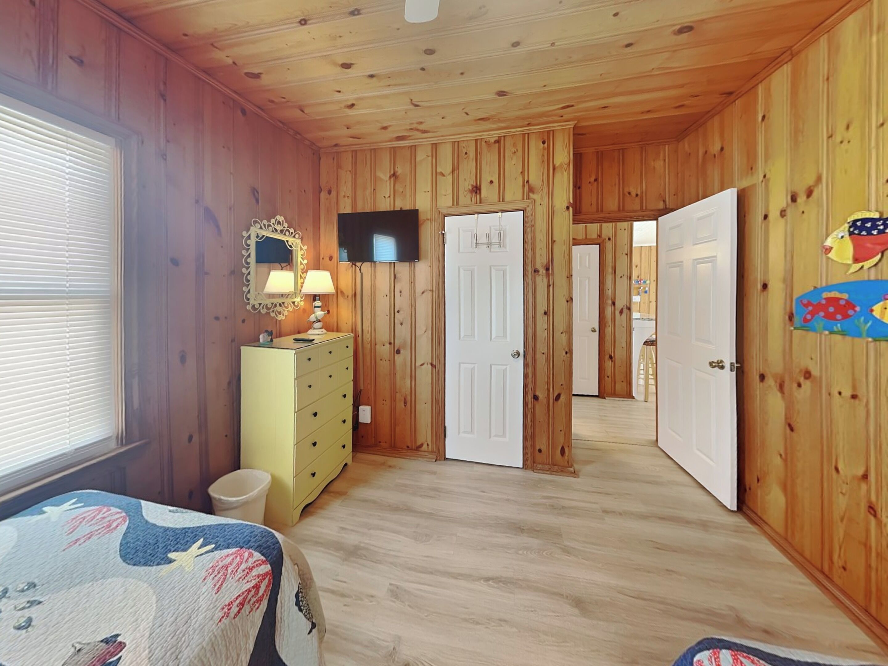 A room with wooden walls and ceiling, a bed with a colorful bedspread, a dresser with a mirror and lamps, and a television on the wall.