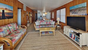 Ein gemütliches Wohnzimmer mit Sofa, Couchtisch und TV-Schrank. Holzverkleidungen an den Wänden und eine Küche im Hintergrund.