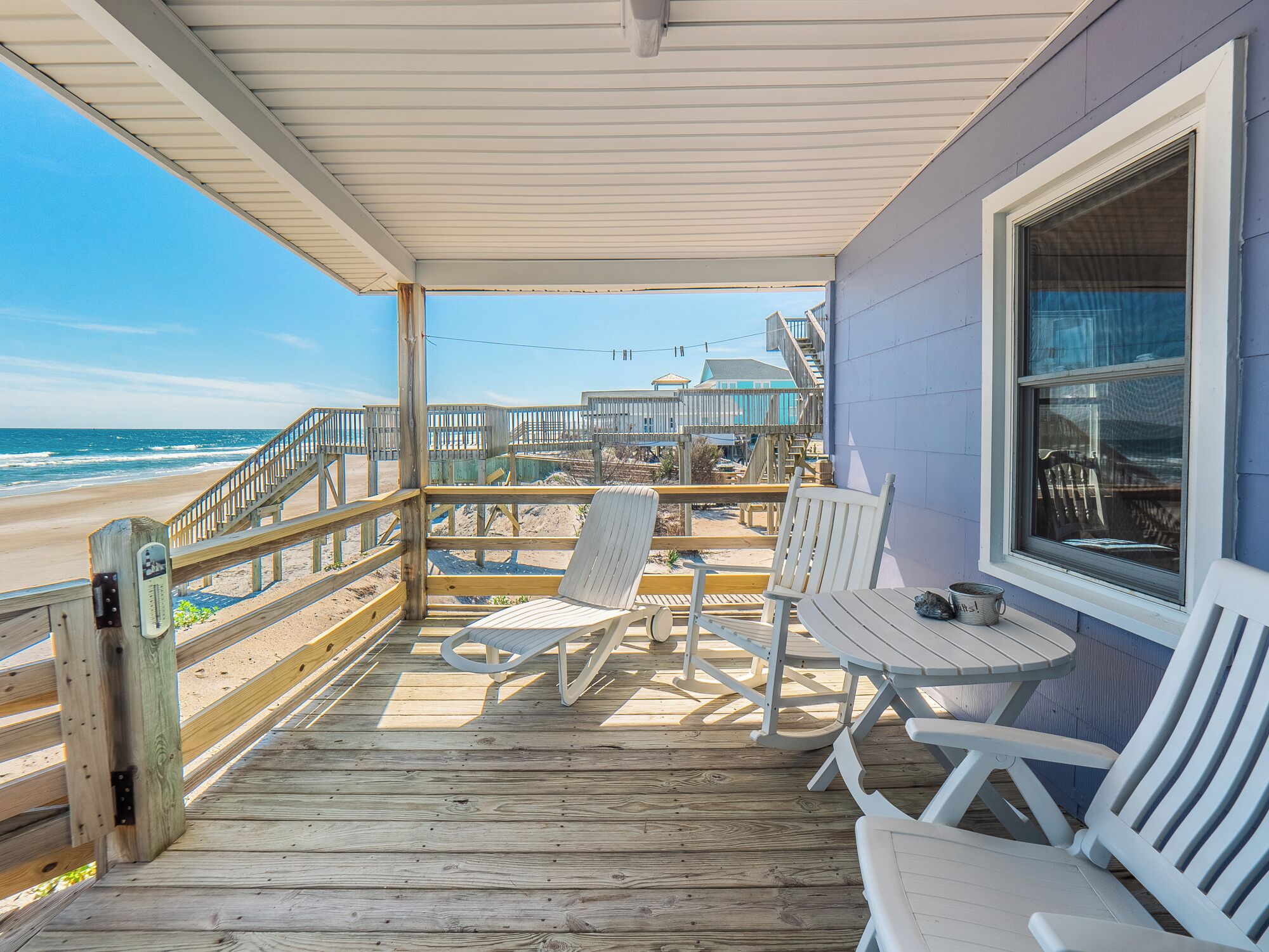 A wooden deck with white chairs and a table, overlooking a beach and a pier.
