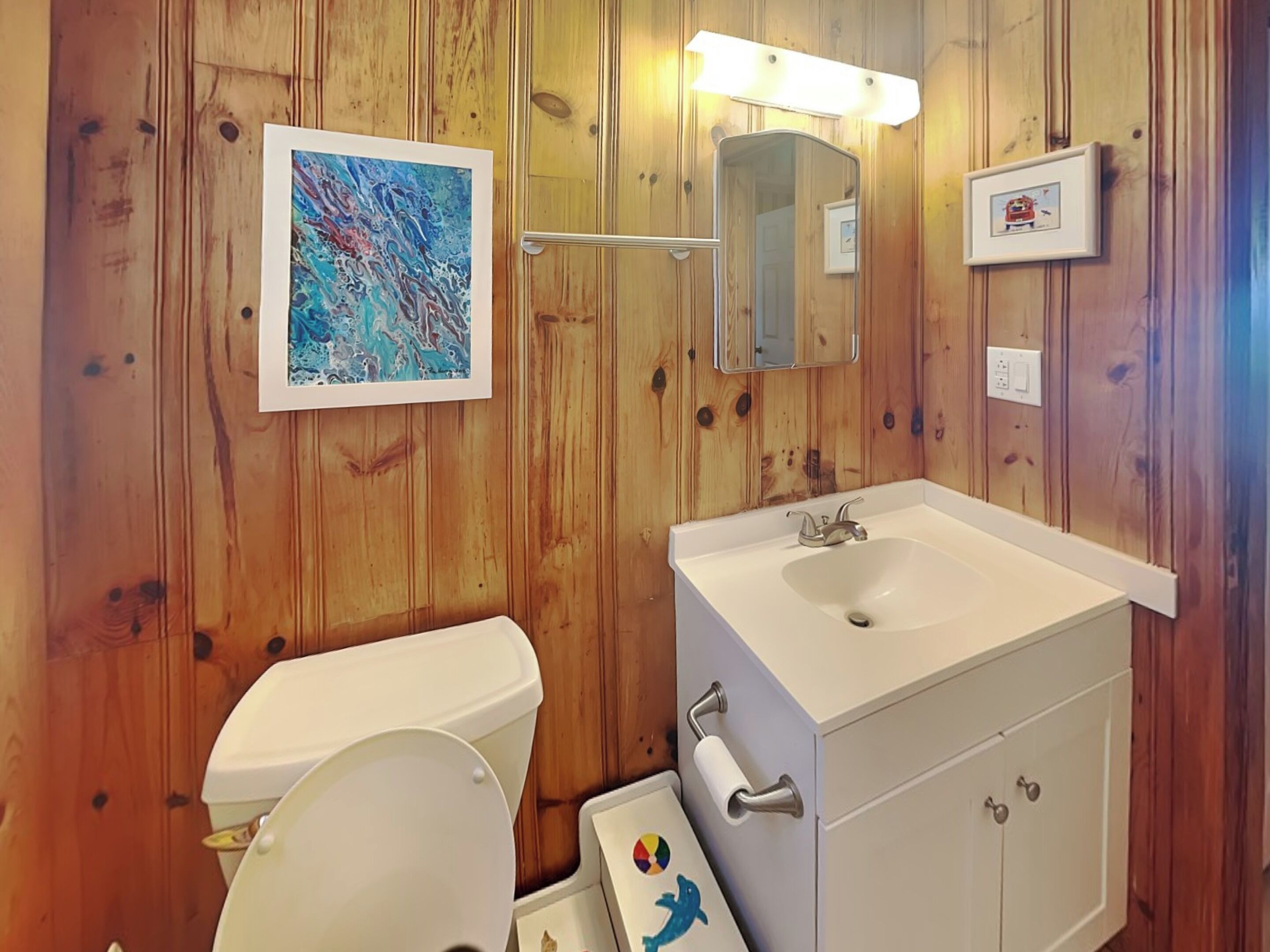 Une salle de bain avec un lambris en bois, un lavabo blanc, un miroir et un tableau abstrait encadré.