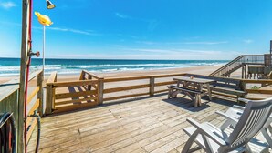 Une terrasse en bois avec un banc et une table, donnant sur une plage où l’on peut admirer les vagues.